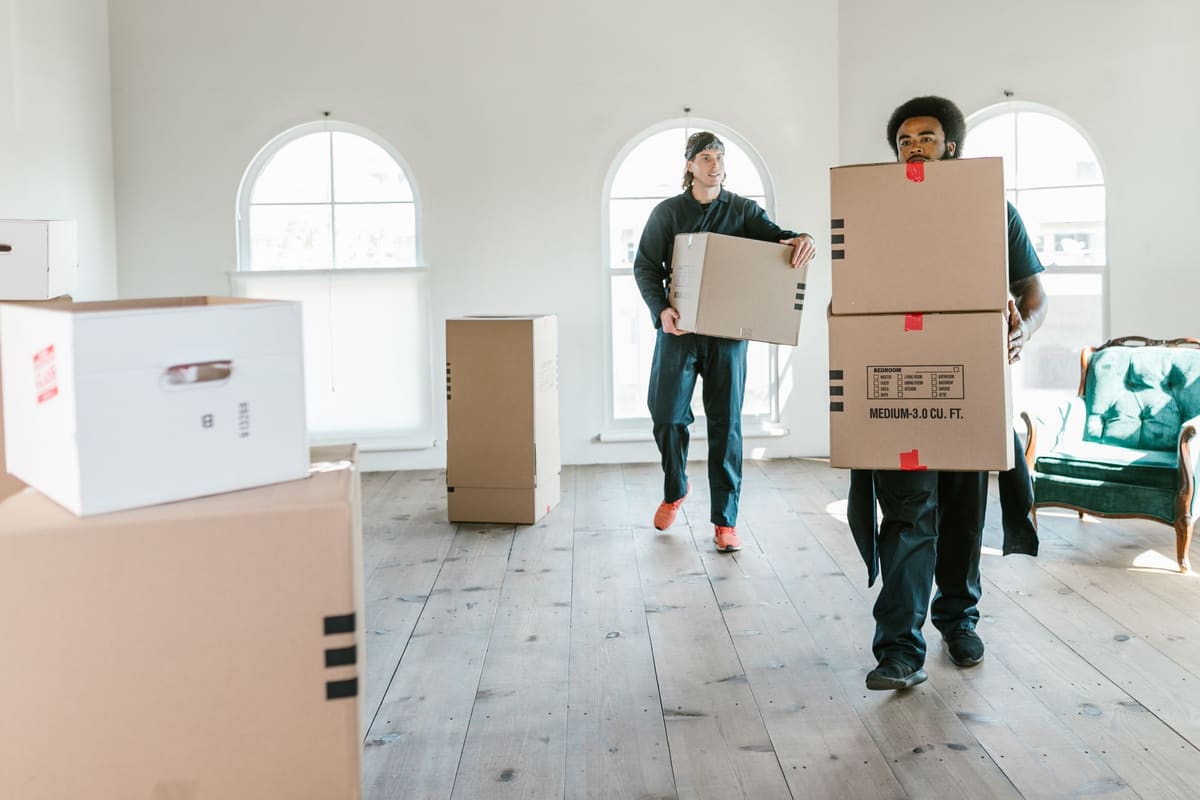 Two people carrying moving boxes through an empty room with boxes stacked along the wall