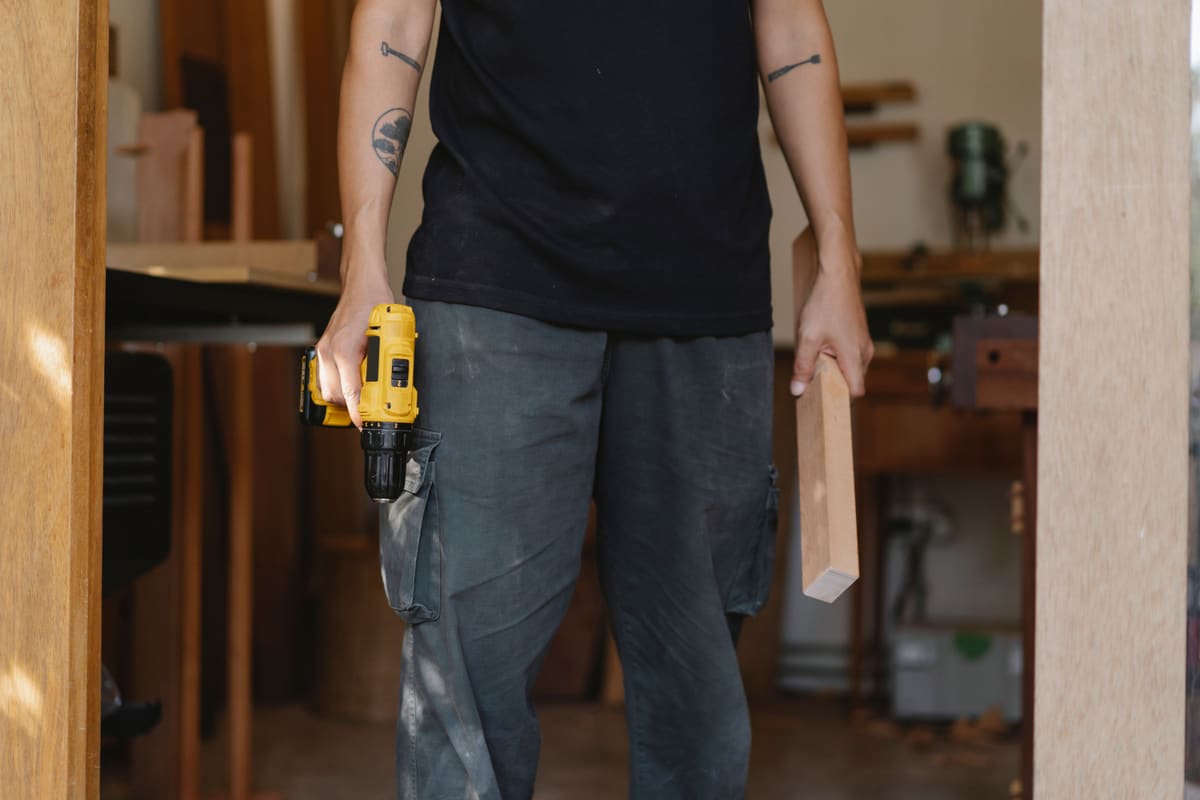 Tradesperson carrying a cordless drill and a length of timber through a workshop
